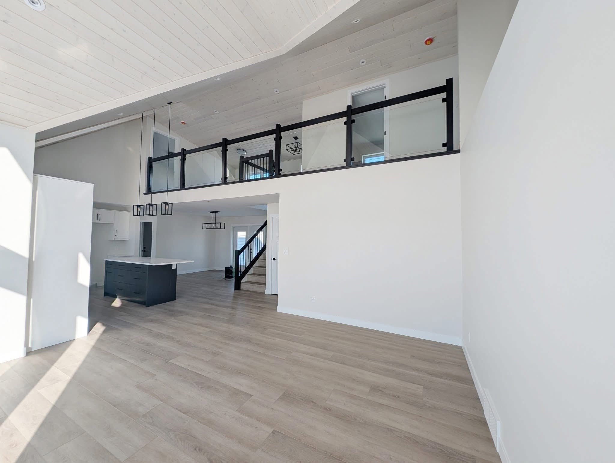 Spacious modern interior with high ceiling and wooden floor. Features an open loft with glass railing, kitchen island, and large windows casting light.