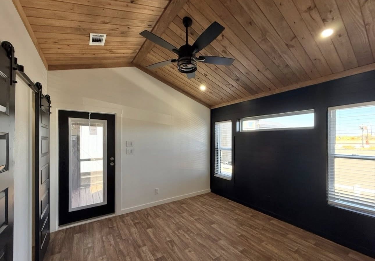 Cozy room with wood-paneled sloped ceiling, black ceiling fan, and floor-to-ceiling windows. Light wood flooring and a black-framed glass door.