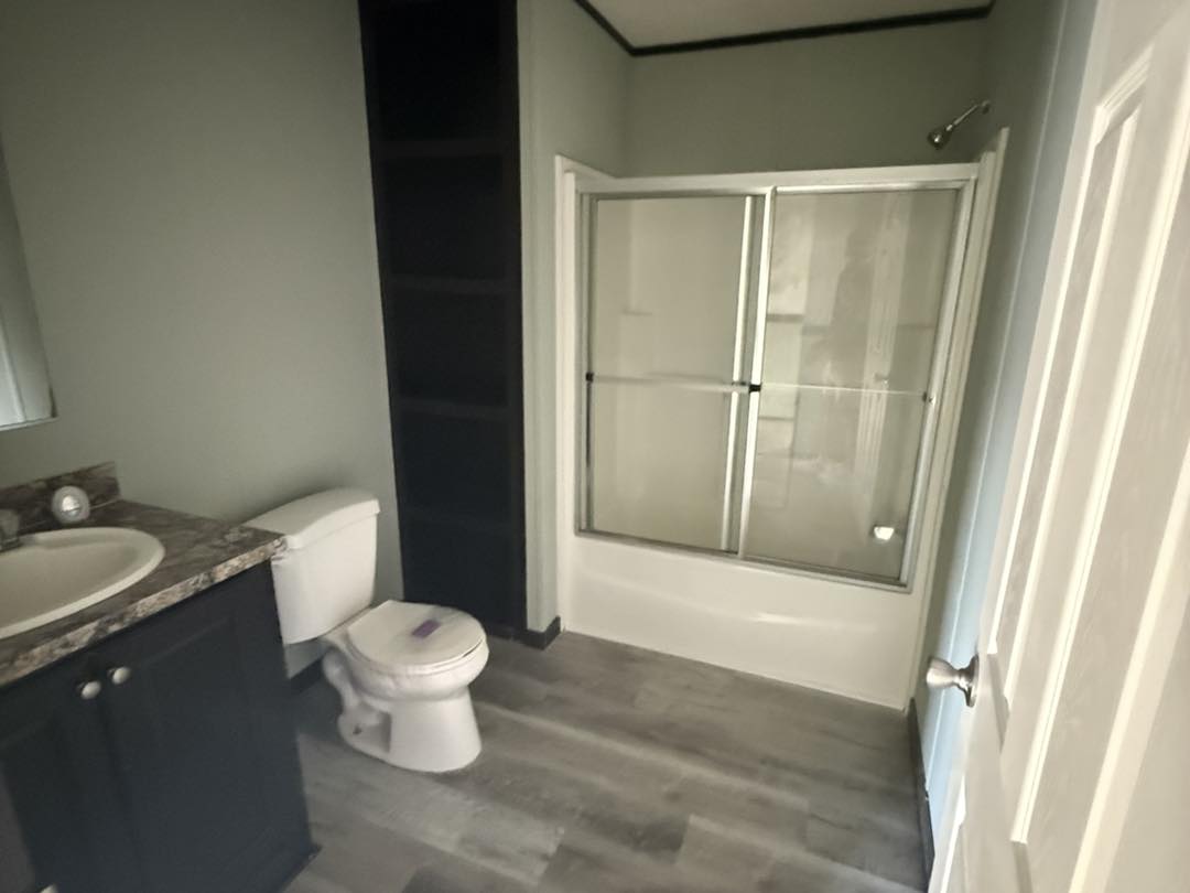 Modern bathroom with gray wood flooring, featuring a dark vanity with granite countertop, toilet, and a glass-enclosed bathtub shower. Neutral tones create a calm ambiance.