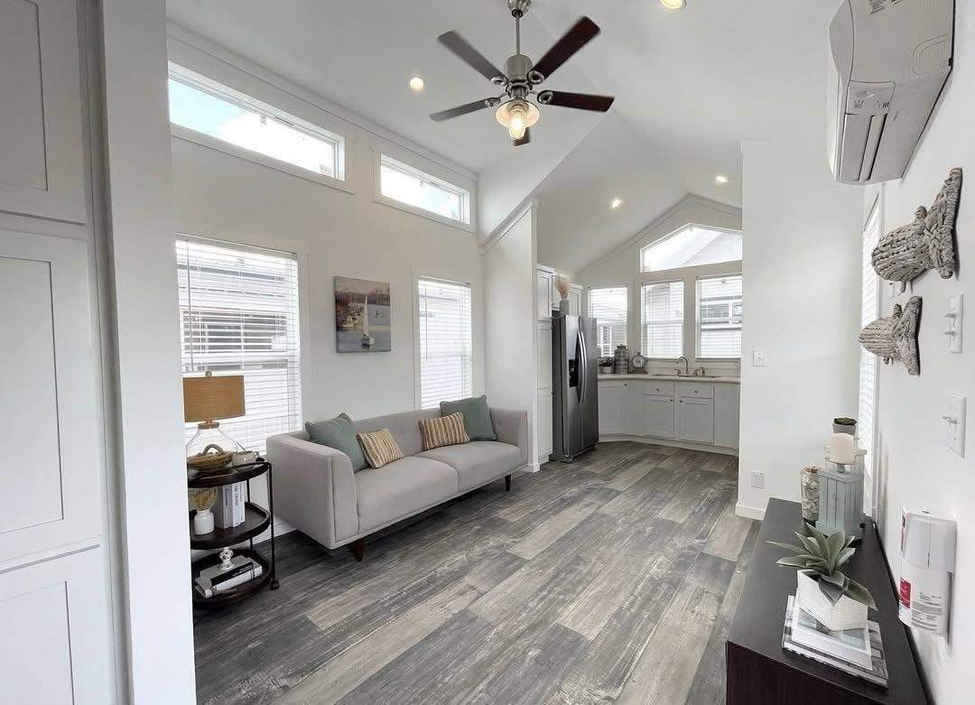 Modern, airy living room with vaulted ceilings, a gray sofa, and wood-look flooring. Kitchen area with silver appliances. Minimalist decor and a ceiling fan.