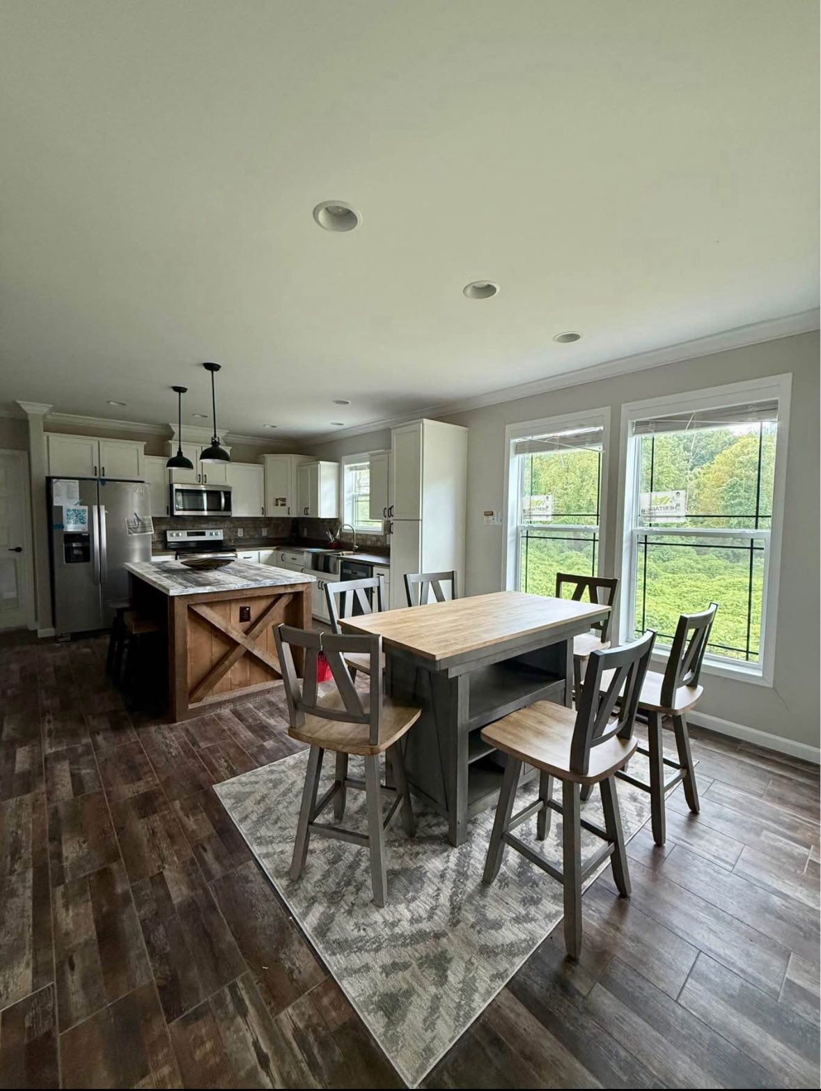 Modern kitchen with wooden flooring, large windows, and a kitchen island. Features a wooden dining table, chairs, and pendant lights, evoking a cozy, open atmosphere.