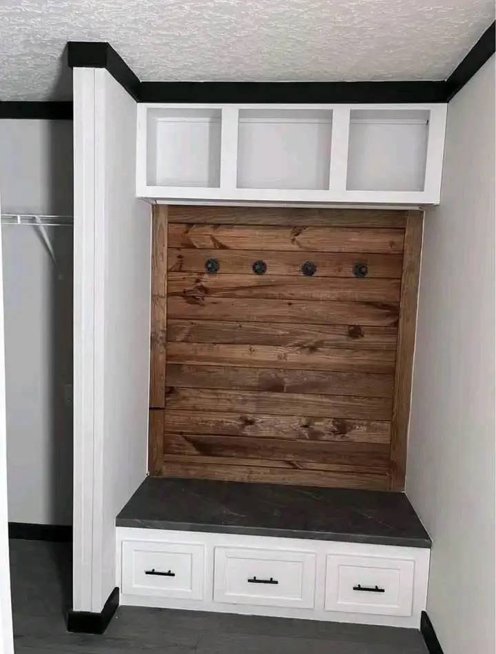 Mudroom area with a wooden accent wall and four black hooks. There’s a gray bench below and white cabinets above for storage, creating a cozy, rustic vibe.