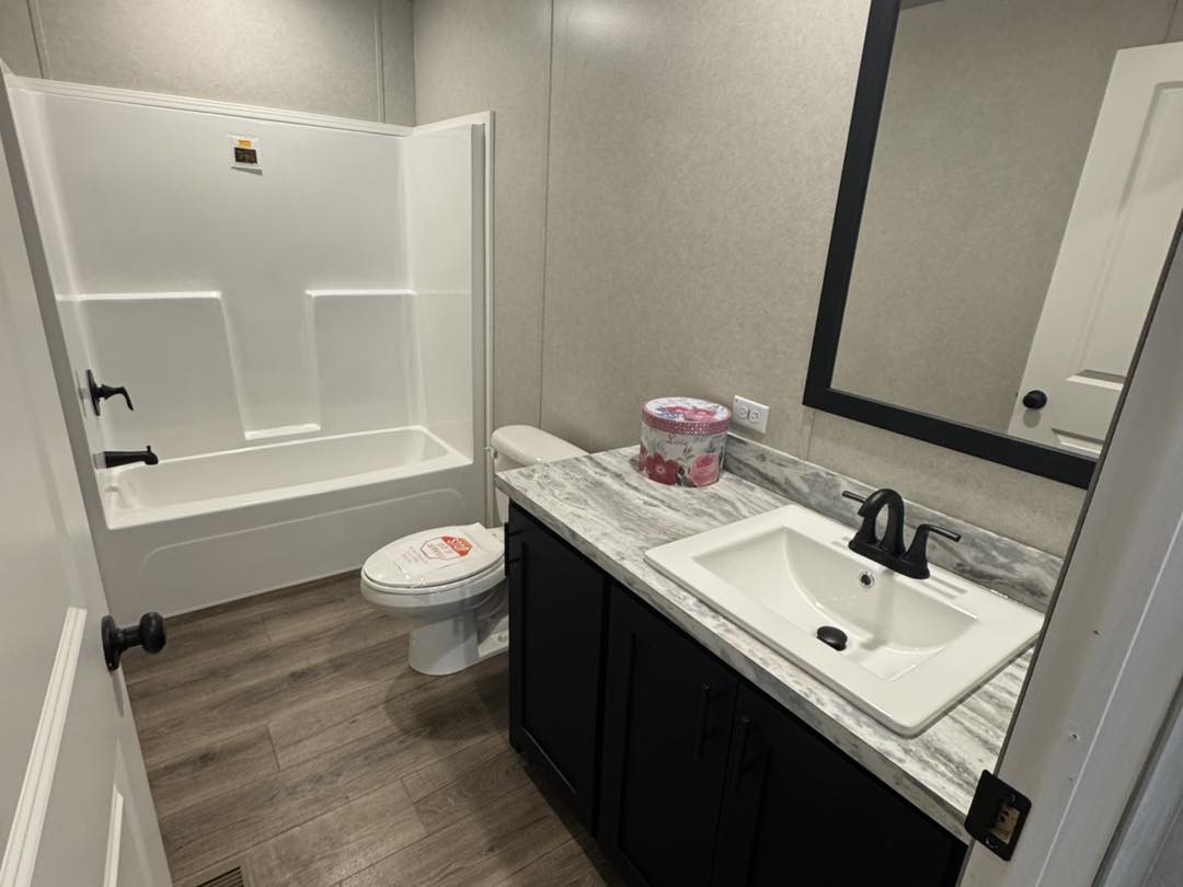 Modern bathroom with a white tub-shower combo, marbled countertop, black fixtures, sink, and storage below. Grey walls and wood-style flooring. Clean and simple.