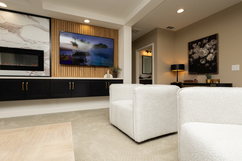 A modern living room with white chairs, a wall-mounted TV displaying a scenic view, a black cabinet, and abstract artwork. Warm lighting creates a cozy ambiance.