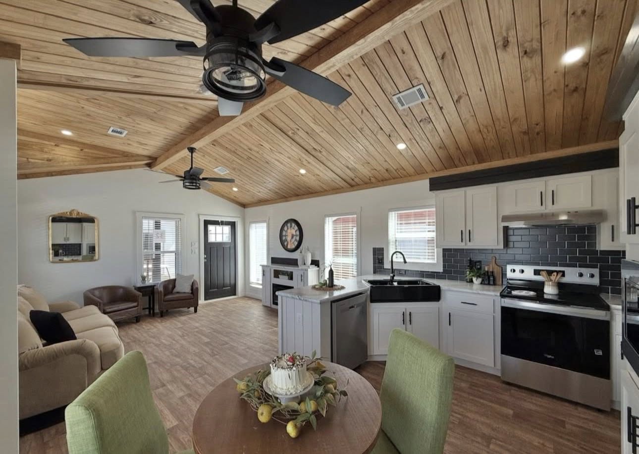 Cozy living room and kitchen with wooden ceiling, modern appliances, round dining table with fruit centerpiece, and a ceiling fan. Bright and inviting space.