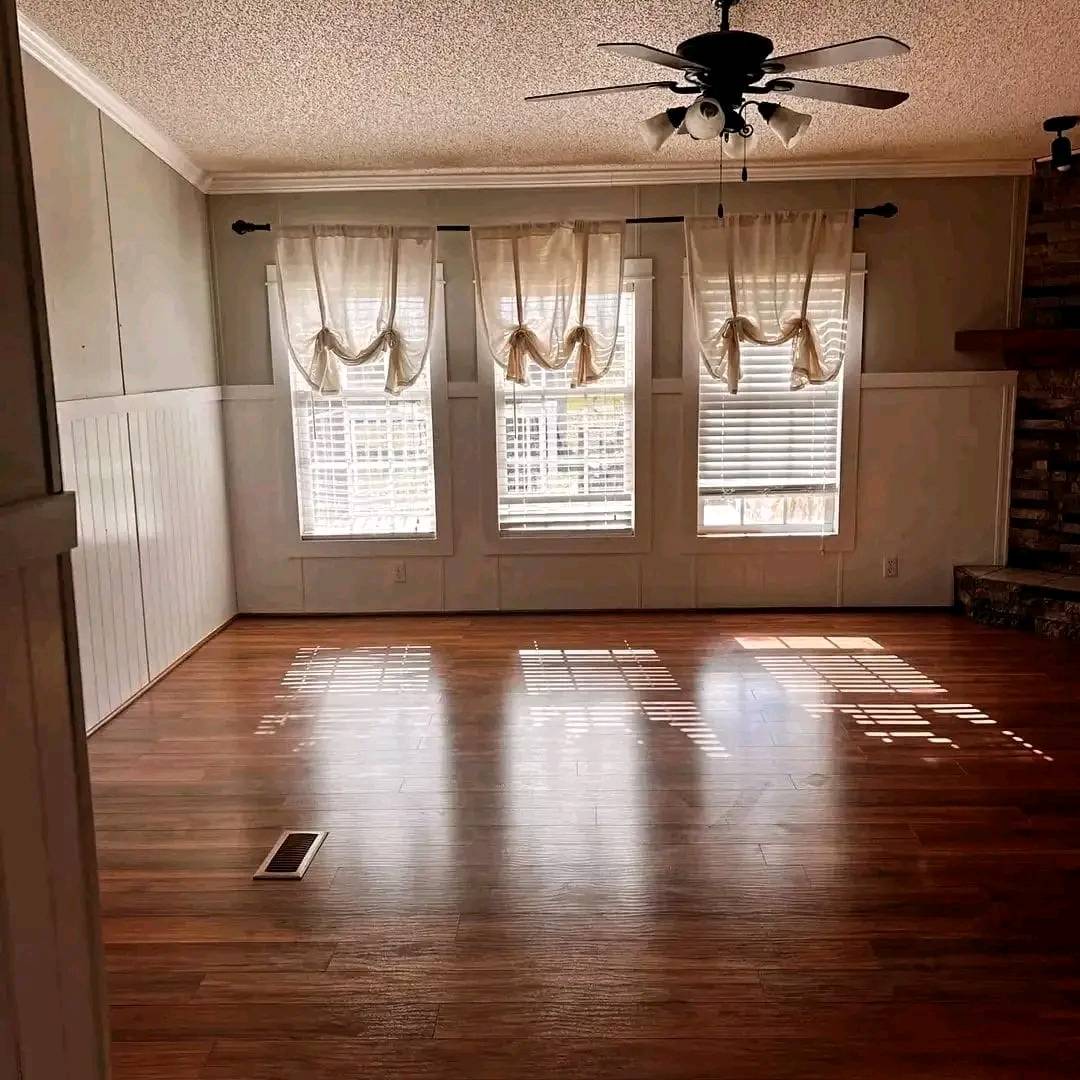 Empty room with wooden floors, three tall windows with sheer curtains, and a ceiling fan above. The sunlight creates striped shadows across the floor, conveying a warm and serene ambiance.