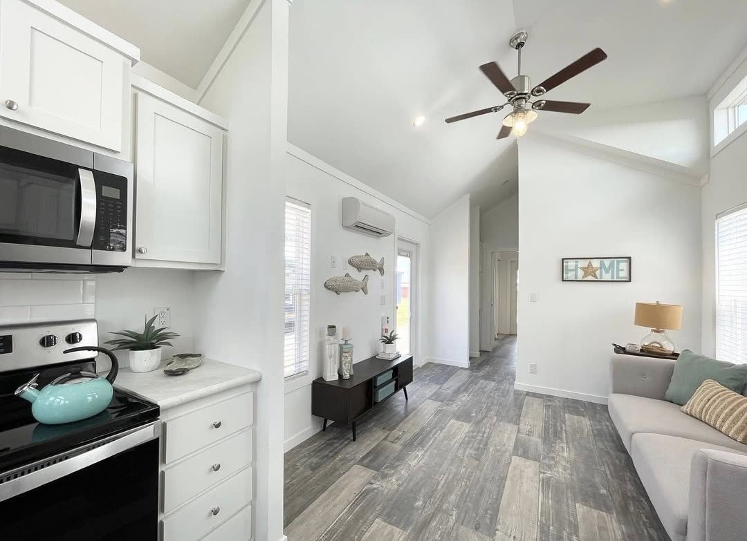 Bright, modern living space with high ceilings and wood-style flooring. A kitchen features white cabinets and stainless appliances. Cozy sitting area with a sofa, lamp, and "Home" wall decor, creating a welcoming tone. Ceiling fan above.