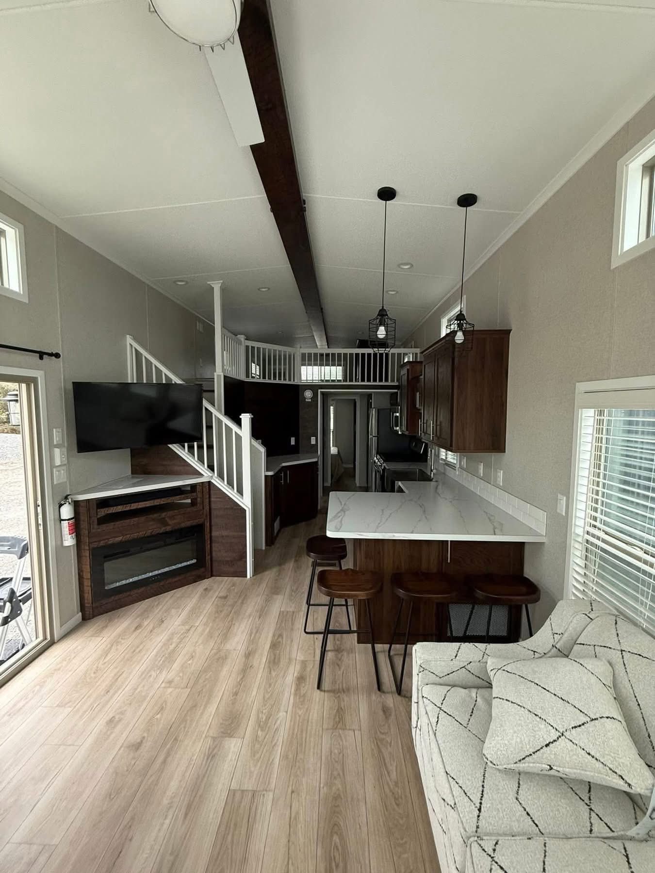 Modern tiny home interior with light wood flooring, loft area, dark wood cabinets, white counters, kitchen island with stools, and cozy patterned sofa.