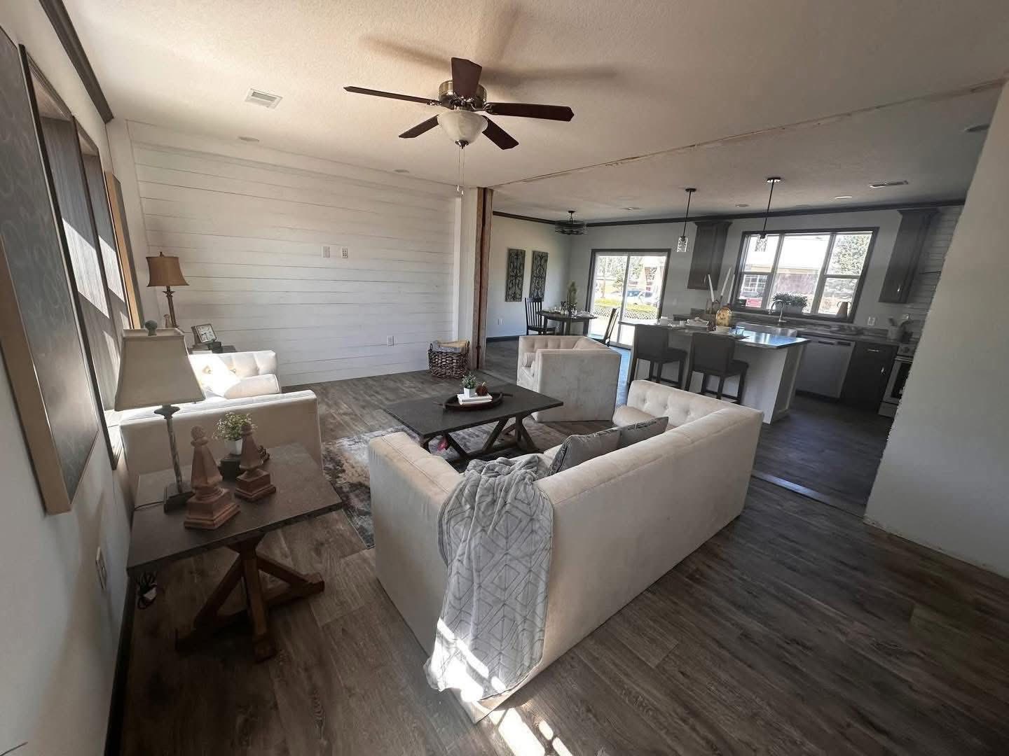 Spacious living room with beige couches, wooden floor, and ceiling fan. Open concept leads to a modern kitchen with a large window and island seating.