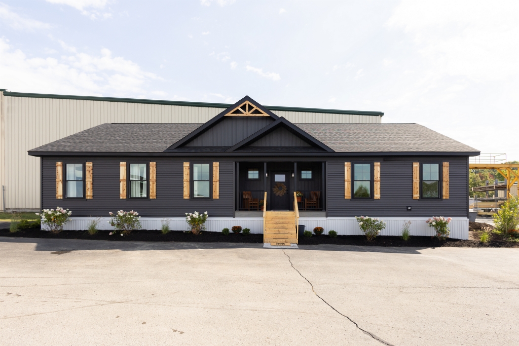 Dark gray modular home with wooden shutters and a small porch. Bright sky, minimal landscaping, and industrial background create a modern yet rustic vibe.