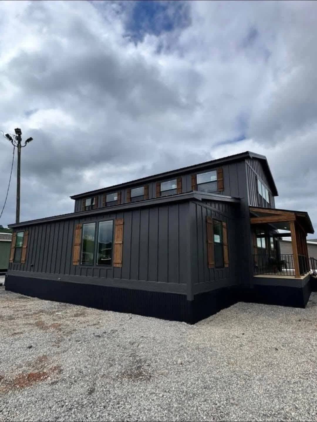 Modern, black tiny house with multiple windows and a wooden awning, under a cloudy sky, situated on a gravel surface, conveying a minimalist vibe.