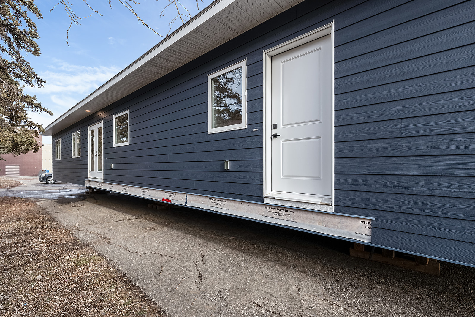 A modular home with dark blue siding is perched on a trailer, showcasing white doors and windows. It rests on a driveway surrounded by trees.