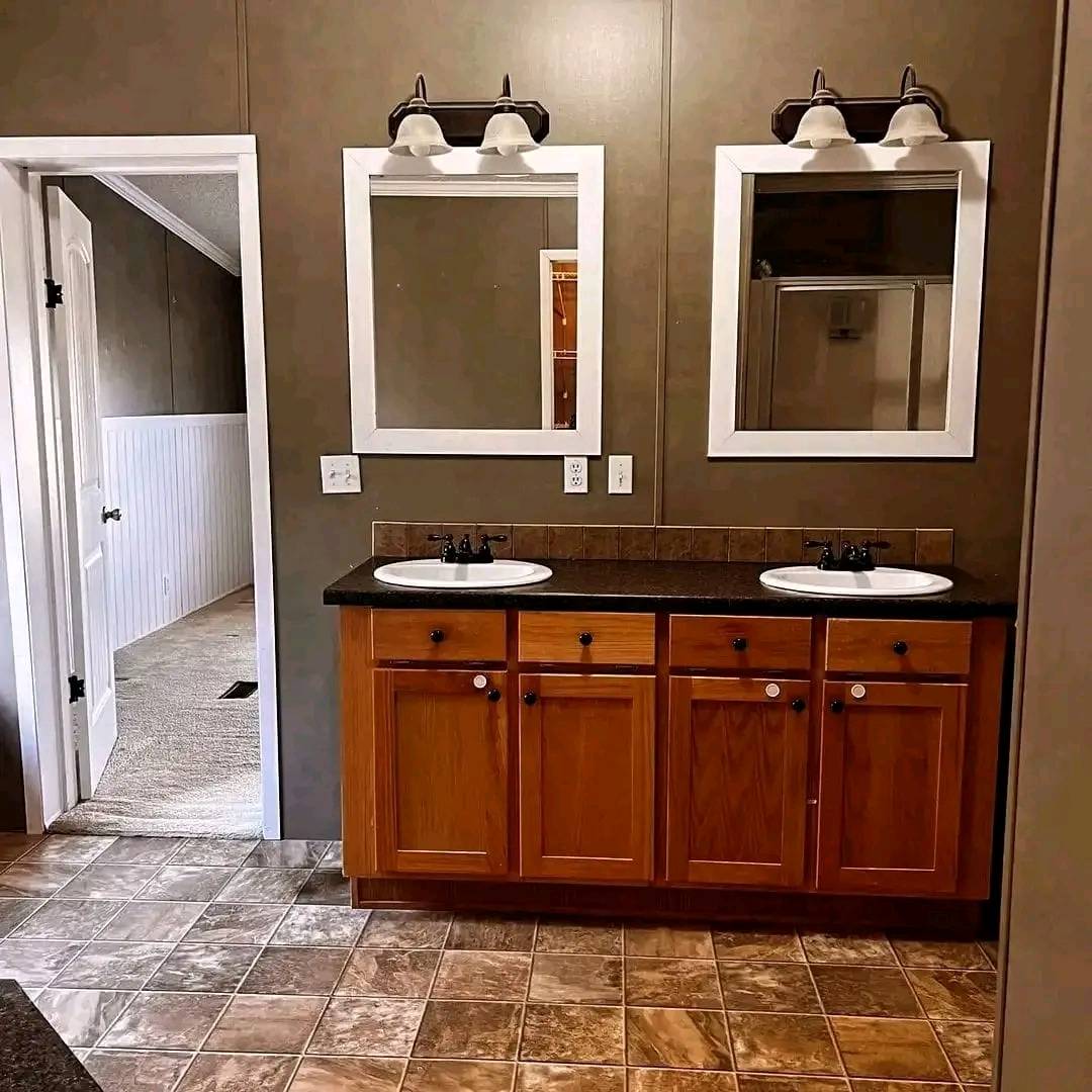 Dual bathroom vanity with wooden cabinets and two white-framed mirrors, under warm lighting. Adjacent doorway leads to a carpeted room.