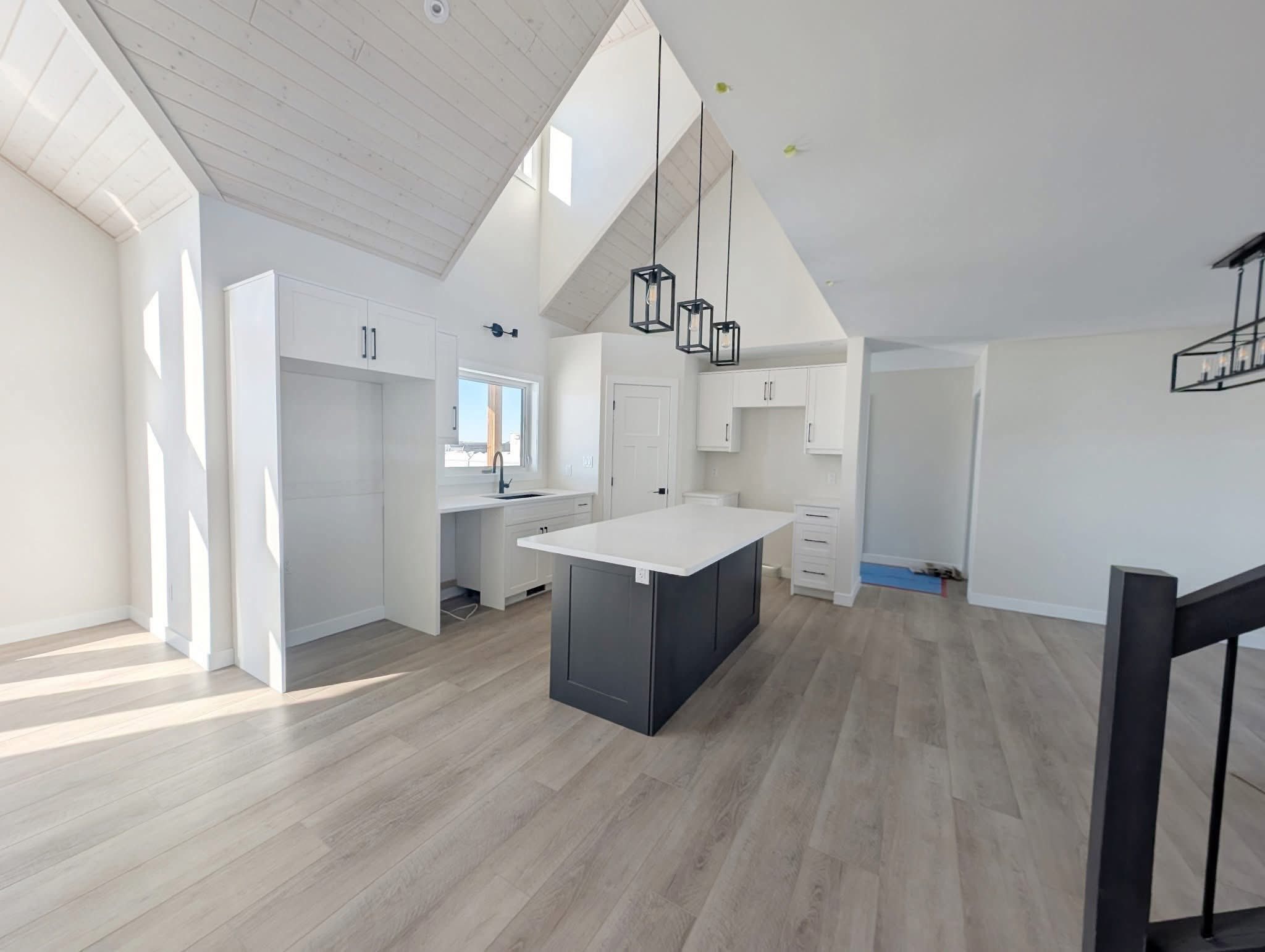 Spacious modern kitchen with high ceilings, featuring white cabinetry, a dark island with white countertop, pendant lights, and light wood flooring.