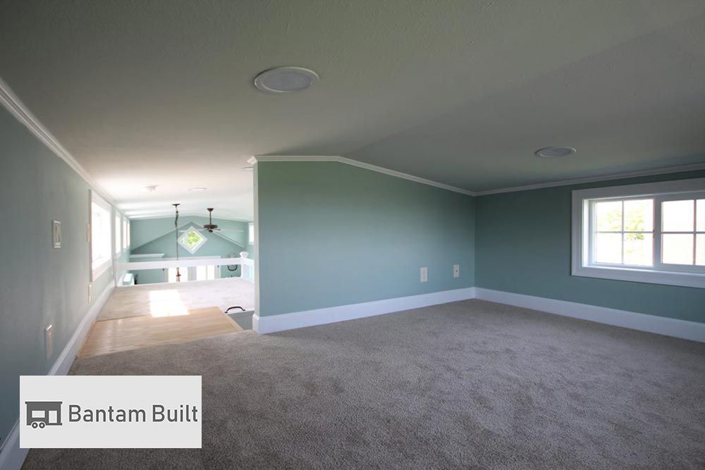 Loft interior with soft green walls, beige carpet, and a single window. Open view to bright lower level. Calm and spacious ambiance. Logo: Bantam Built.