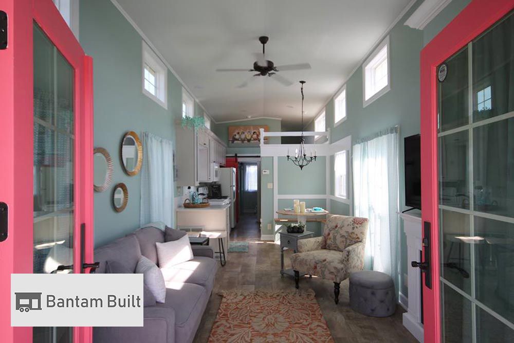 Interior of a small living space with pastel blue walls and red-framed glass doors. Includes a gray sofa, floral armchair, and open kitchen with chandelier, exuding cozy elegance.