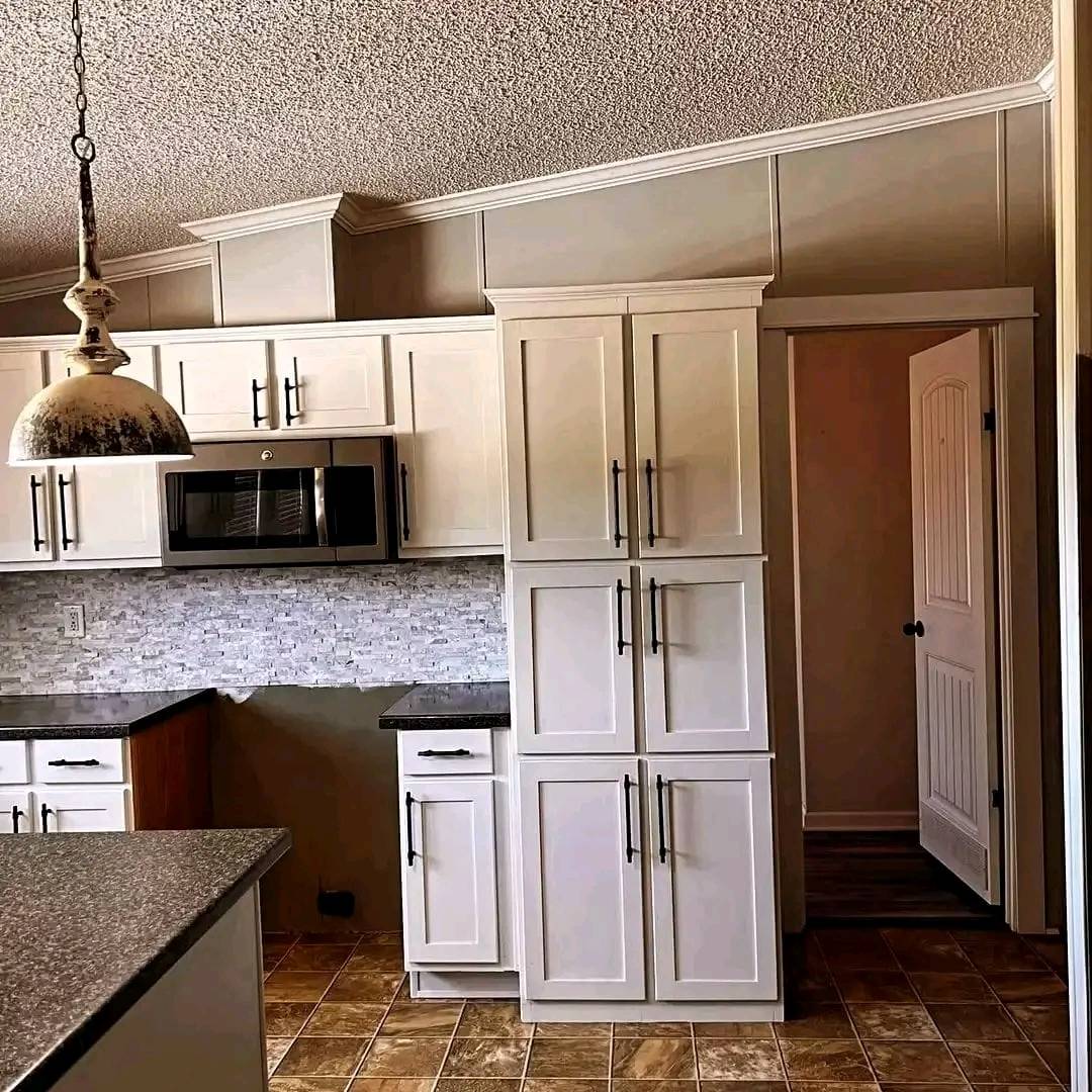 Modern kitchen with white cabinets, black handles, and a built-in microwave. Textured backsplash, gray countertops, pendant light, and tiled floor.