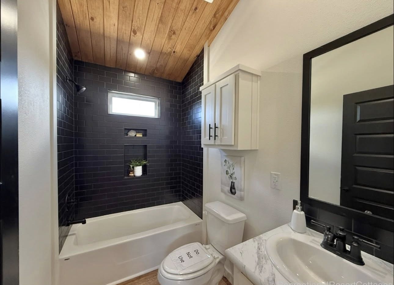 A modern bathroom features a white bathtub with black tile walls and wooden ceiling. A cabinet is above the toilet. The atmosphere is sleek and minimalistic.