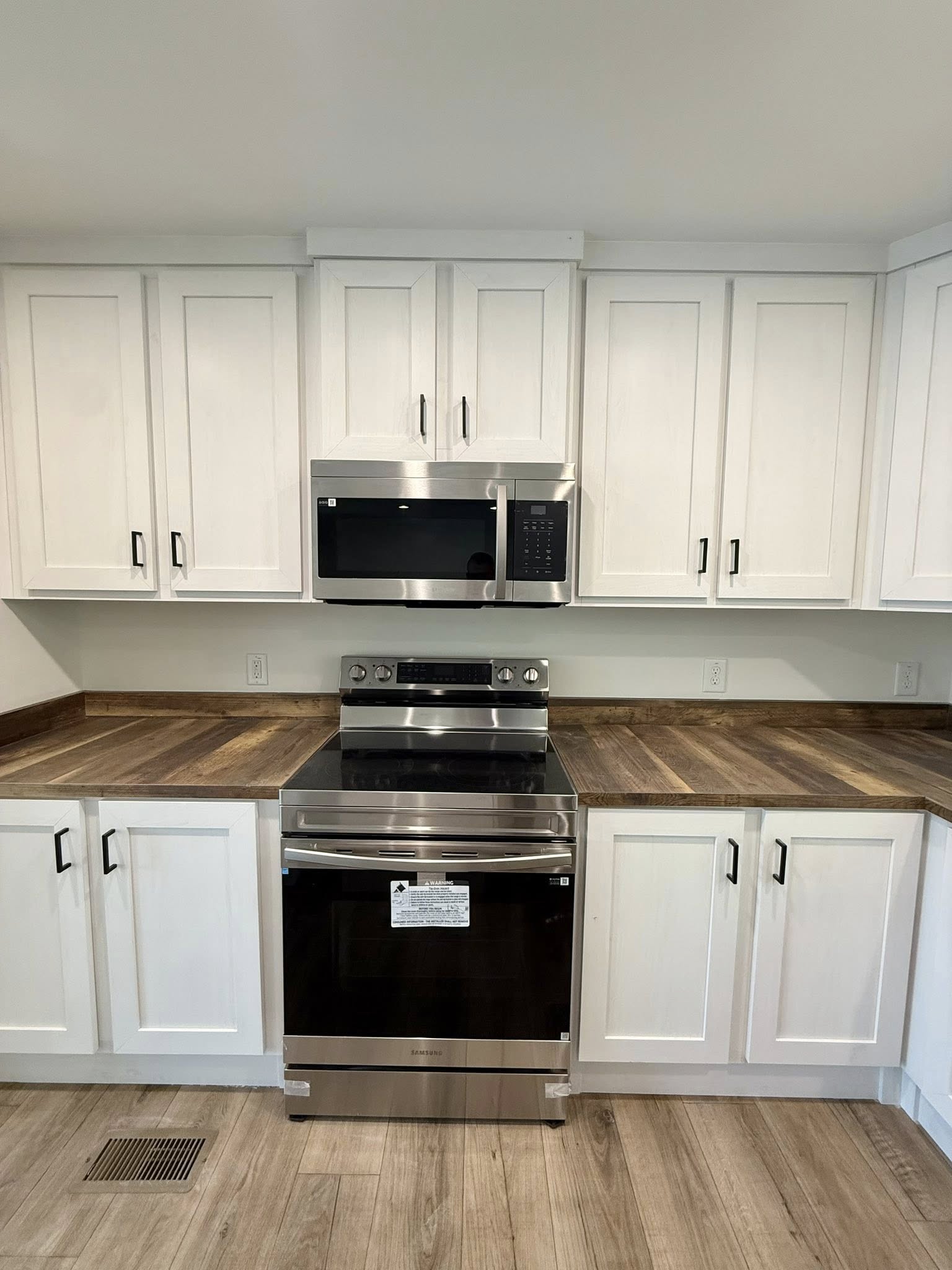 Modern kitchen with white cabinets, stainless steel oven, and microwave. Wooden countertops and flooring add warmth, creating a clean, inviting space.