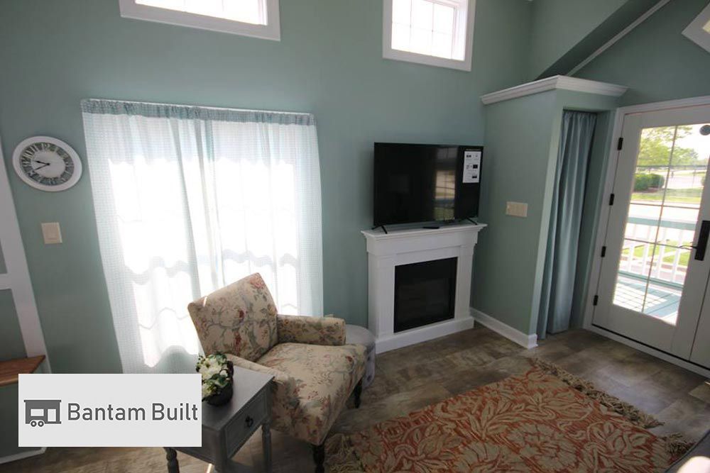 Cozy living room with teal walls, floral armchair, small table, and fireplace topped with a TV. Sunlight streams through sheer curtains, creating a serene atmosphere.