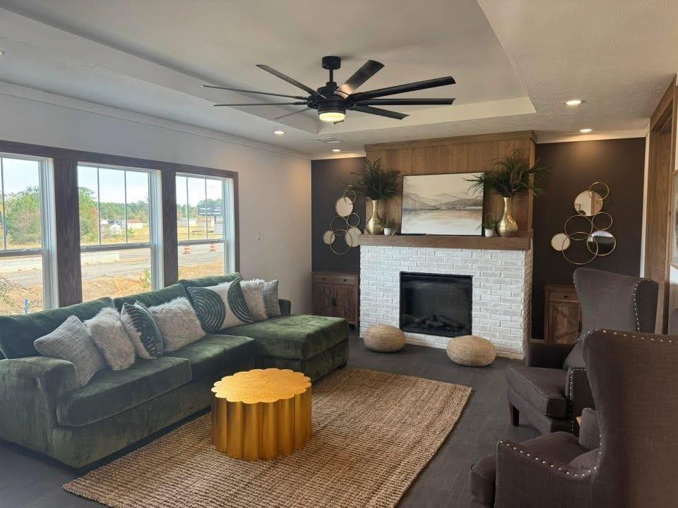 A cozy living room with a green sectional sofa, decorative pillows, and a gold coffee table on a woven rug. The white brick fireplace features plants and art above. Large windows allow natural light, while a ceiling fan adds elegance.