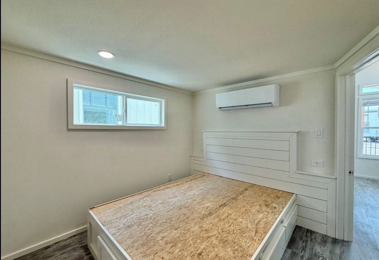 Minimalist bedroom with a white shiplap bed frame, bare plywood mattress, white walls, a narrow window, and an air conditioner. Calm and simple ambiance.