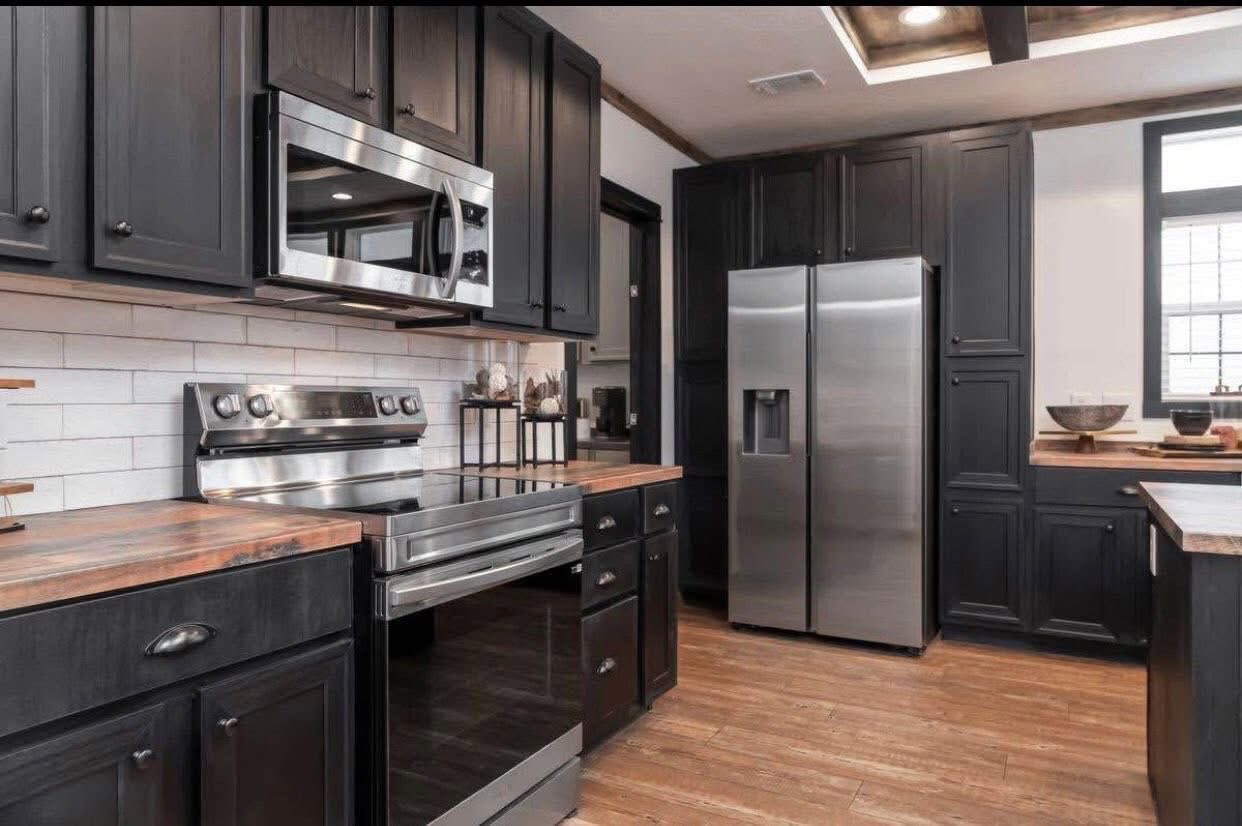 Modern kitchen with dark wood cabinets, stainless steel appliances including a fridge and oven. Light wood floors and a white tile backsplash add contrast.