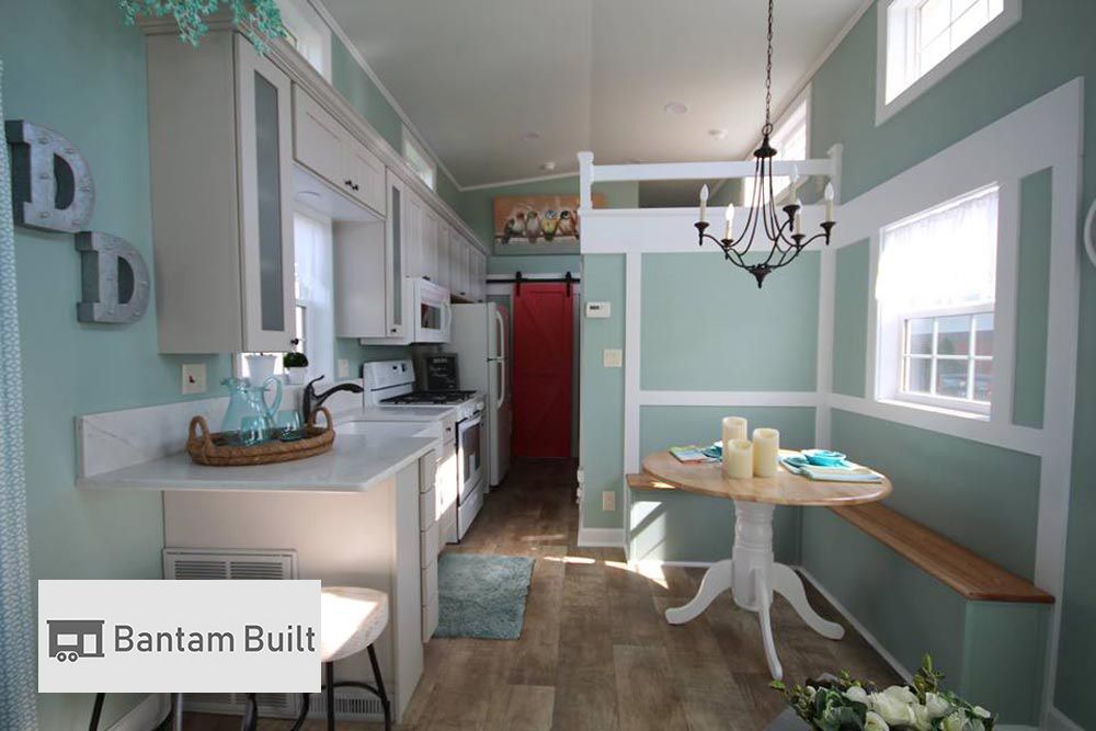 Cozy tiny home interior with mint green walls and wood floor. Features a compact kitchen, red sliding door, round dining table, and chandelier.