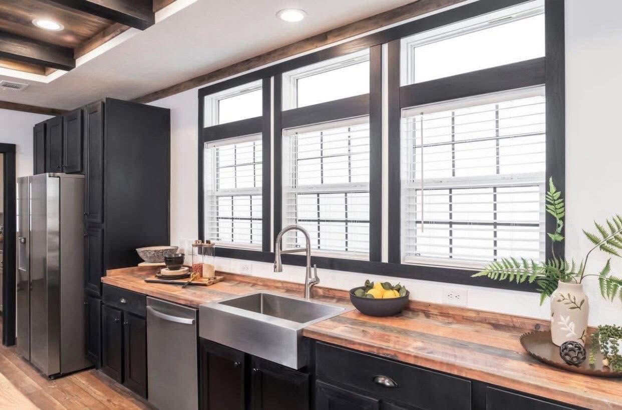 Modern kitchen with black cabinets and wood countertops. Large windows above a stainless steel sink allow natural light. Decor includes fruit and plants.