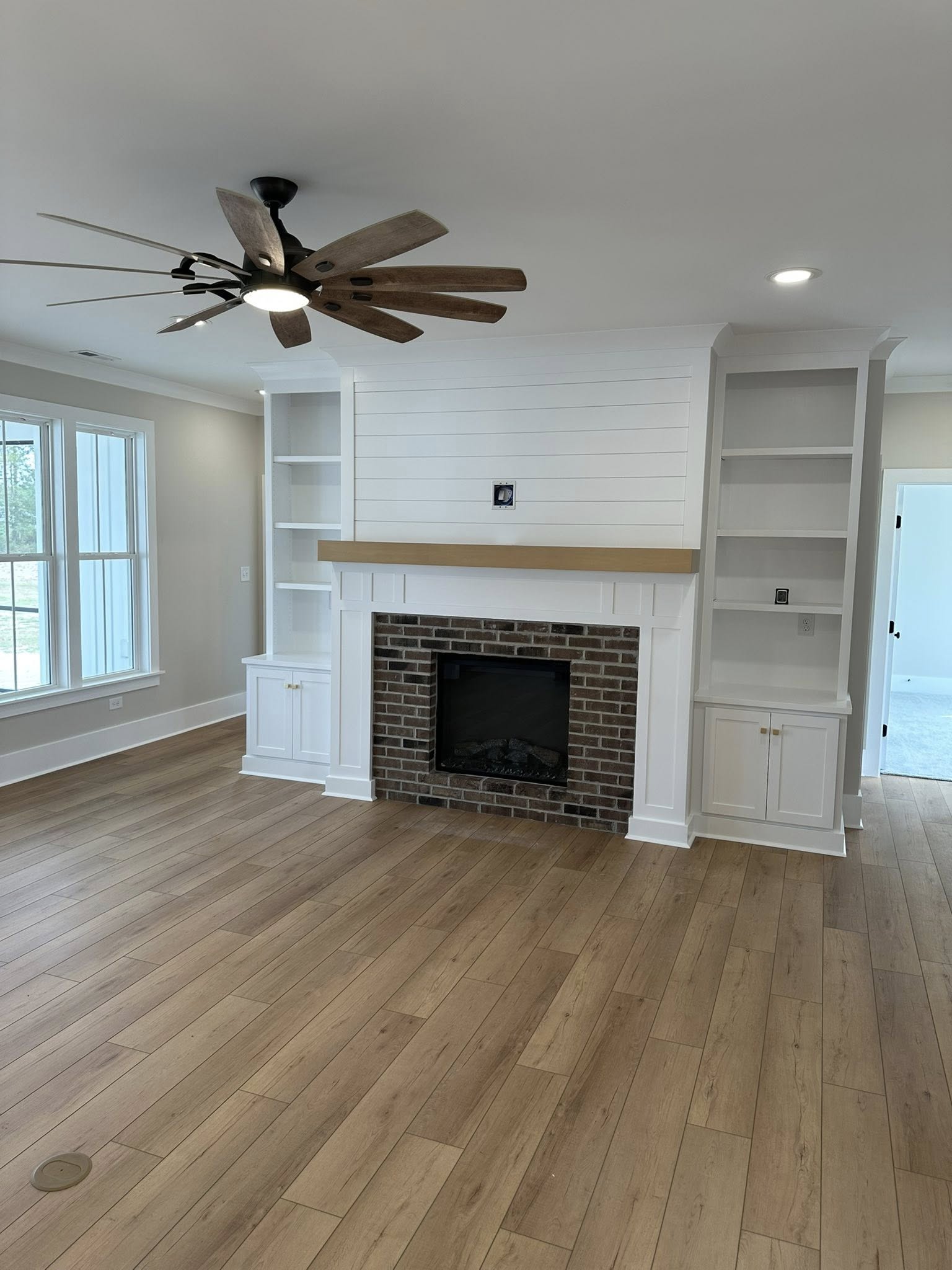 Spacious living room with light wood flooring, a brick fireplace, and white built-in shelves. A modern ceiling fan adds a contemporary touch.