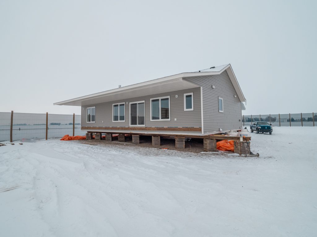 A gray house raised on wooden piers stands on a snowy field. Orange tarps are underneath, with a vehicle in the background. Overcast sky sets a cold tone.