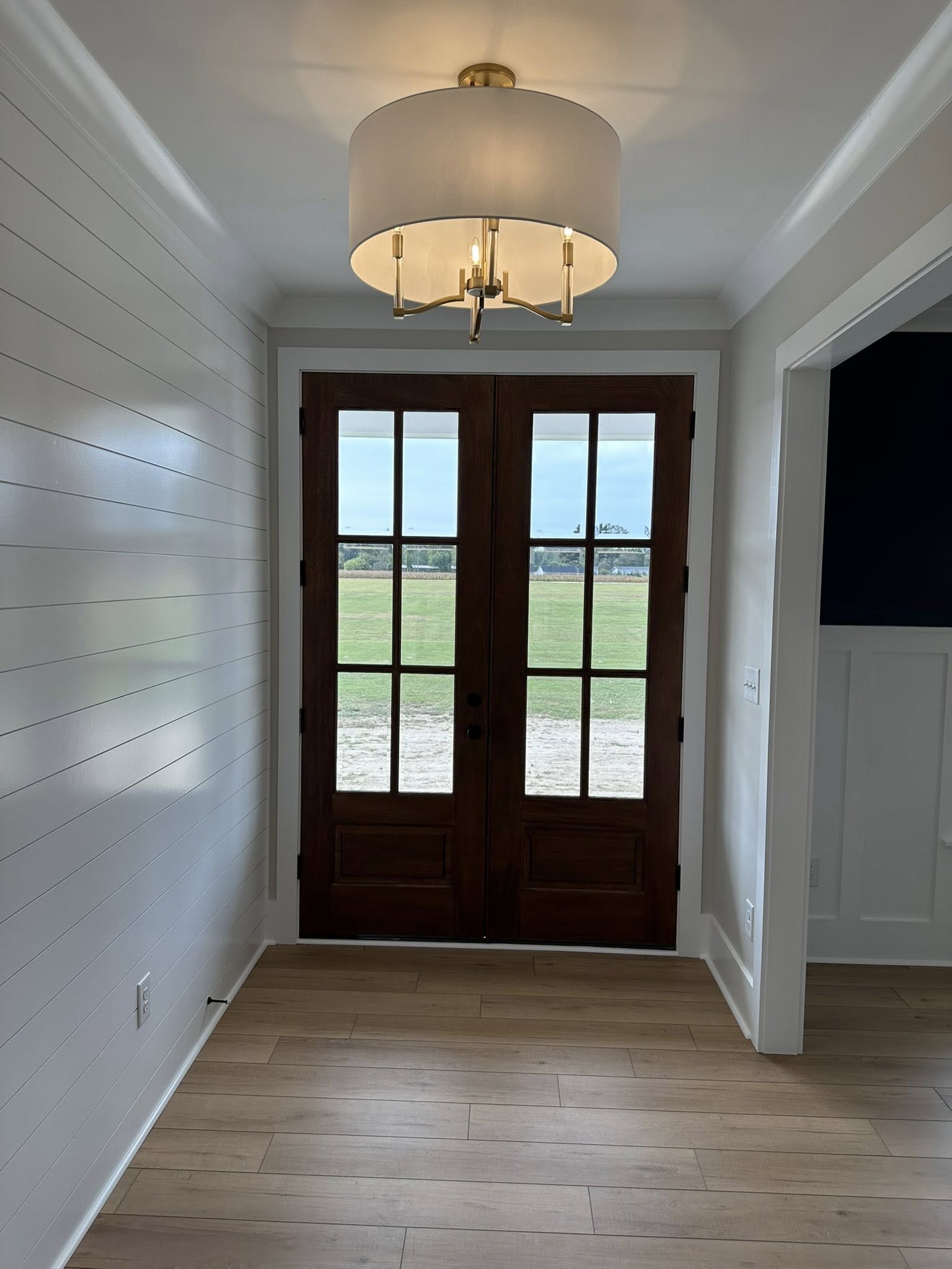 Elegant entryway with double wooden doors featuring glass panes, leading to green fields. A modern chandelier hangs from the ceiling, and light wood flooring complements white walls.