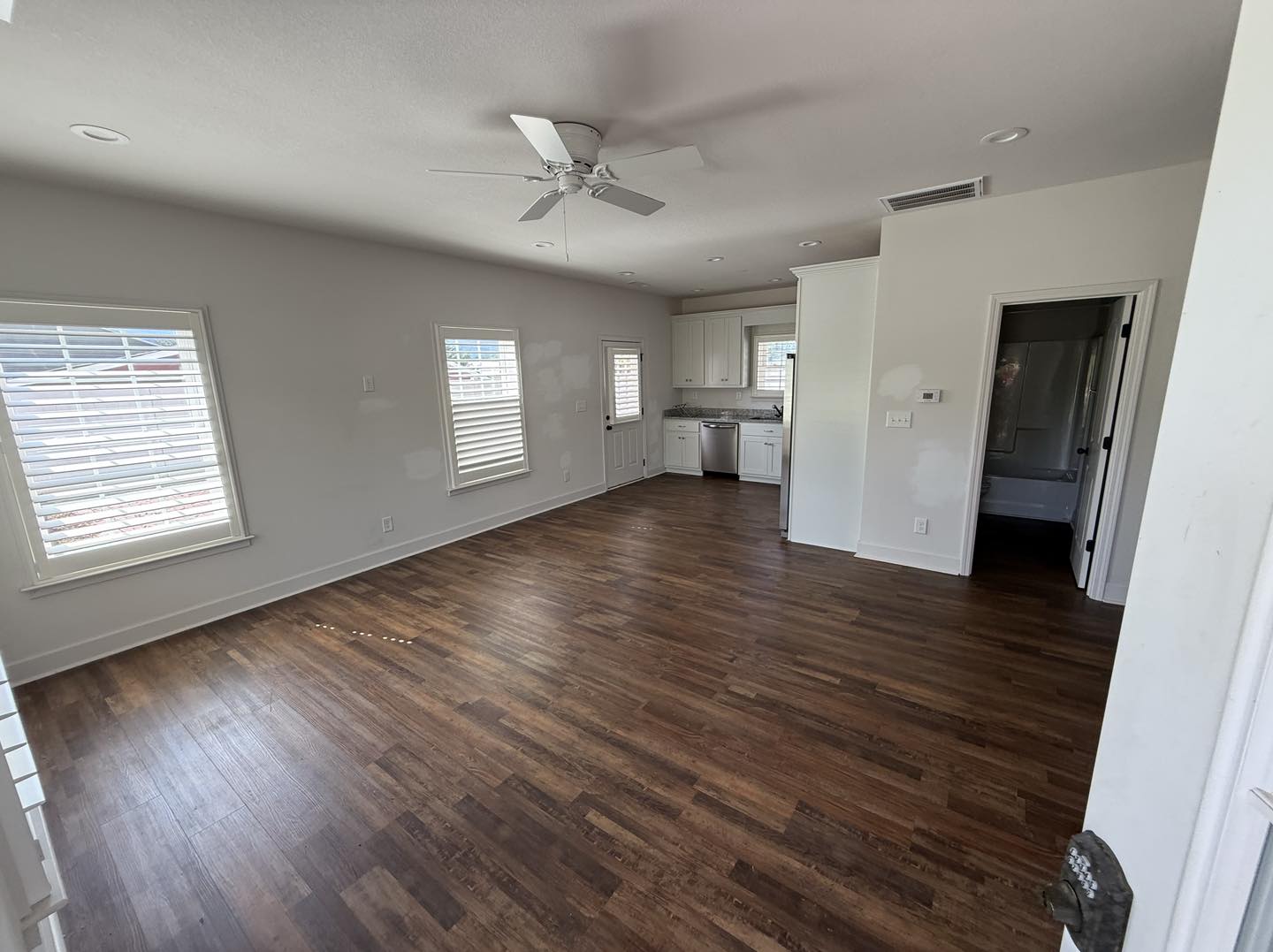 Spacious, empty room with wood flooring and white walls. Windows with blinds let in natural light. Kitchen with white cabinets and door visible.