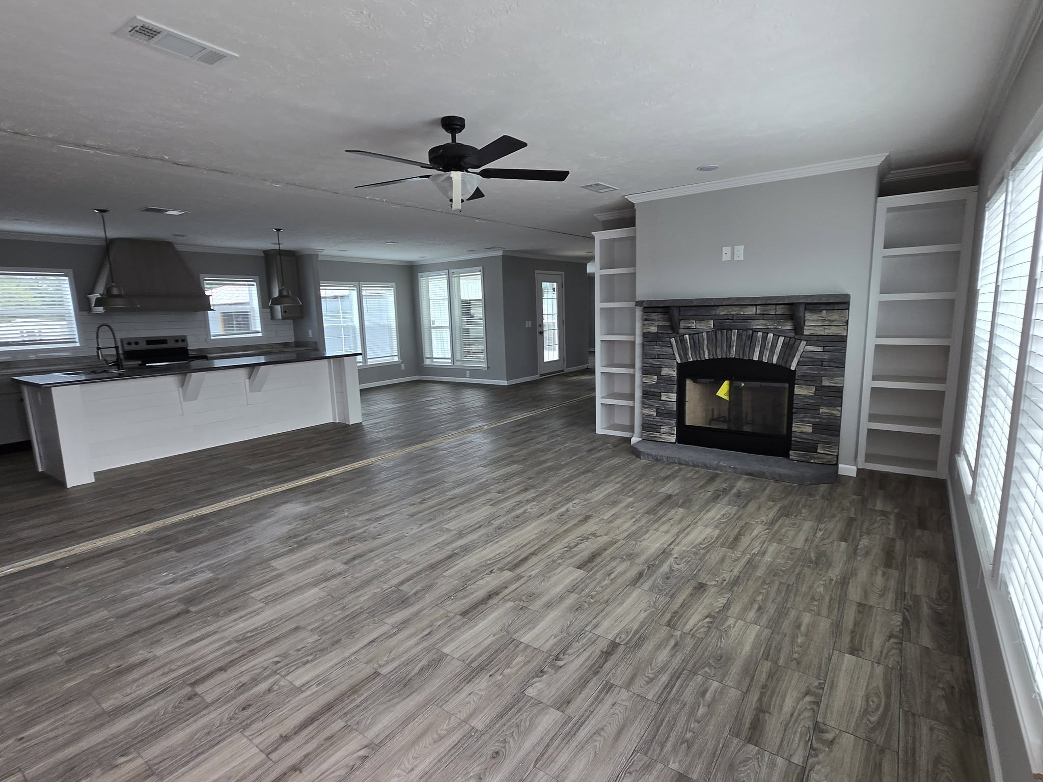 Spacious open-plan living area with wood flooring, a stone fireplace with shelves, large windows, ceiling fan, and adjacent modern kitchen with island.