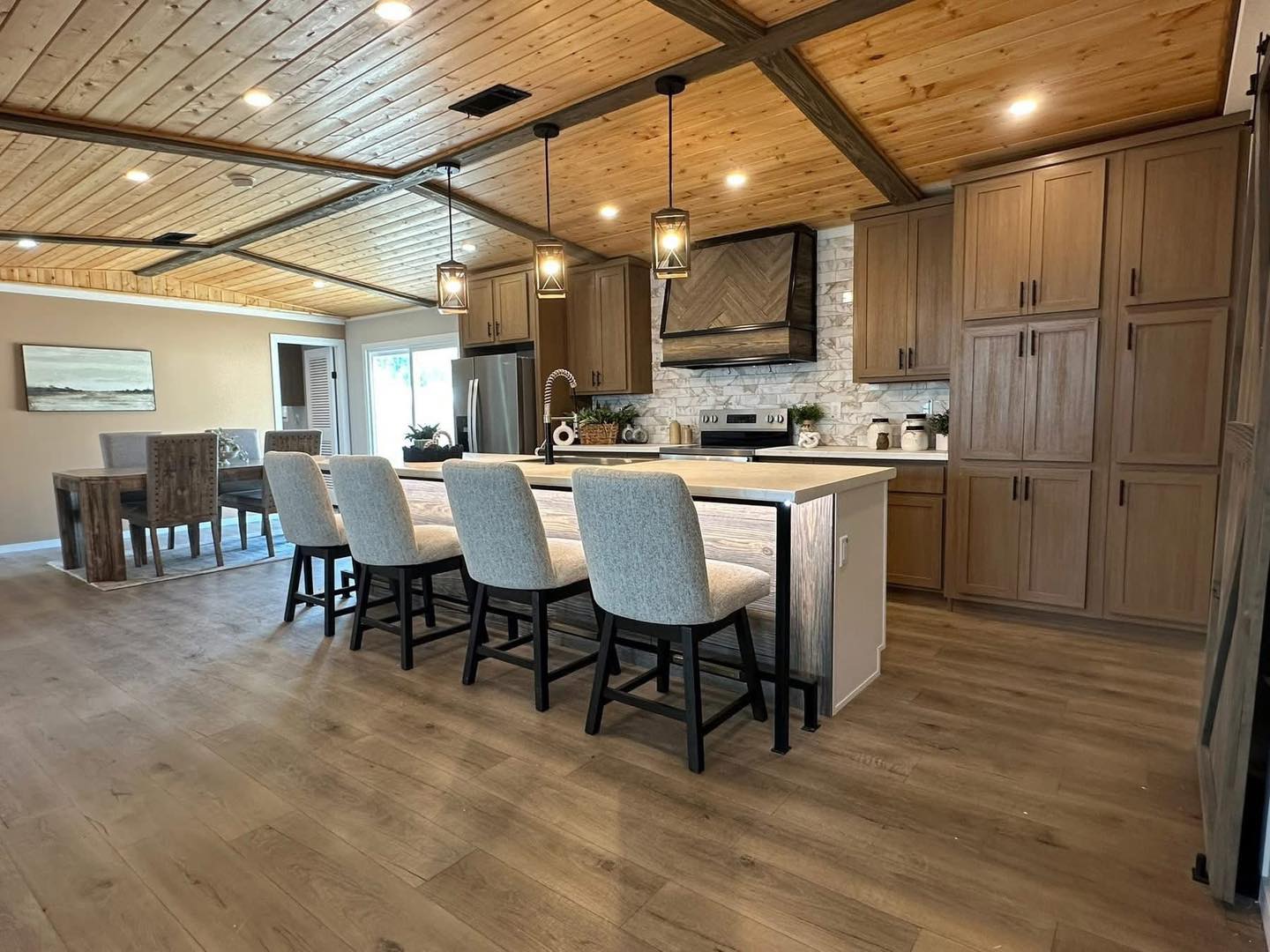 Spacious kitchen with wood-paneled ceiling, light wood floors, and cabinets. An island with four cushioned chairs faces modern appliances, exuding warmth.
