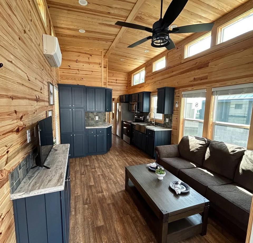 A cozy wooden living room with high ceilings, a dark sofa, and a coffee table. The kitchen features navy cabinets and stainless steel appliances.