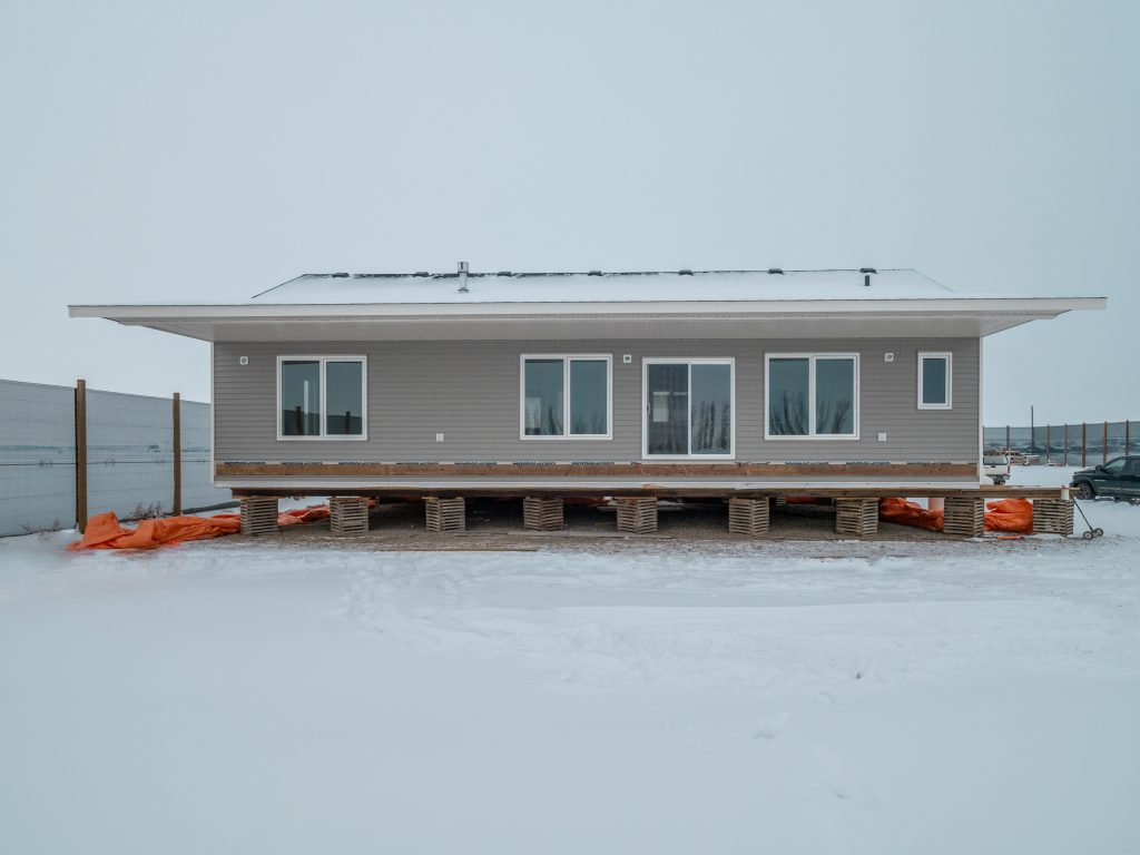 A modern, elevated, grey house on concrete blocks in a snowy landscape. It has large windows, a flat roof, and orange tarps underneath, evoking a cold, serene atmosphere.