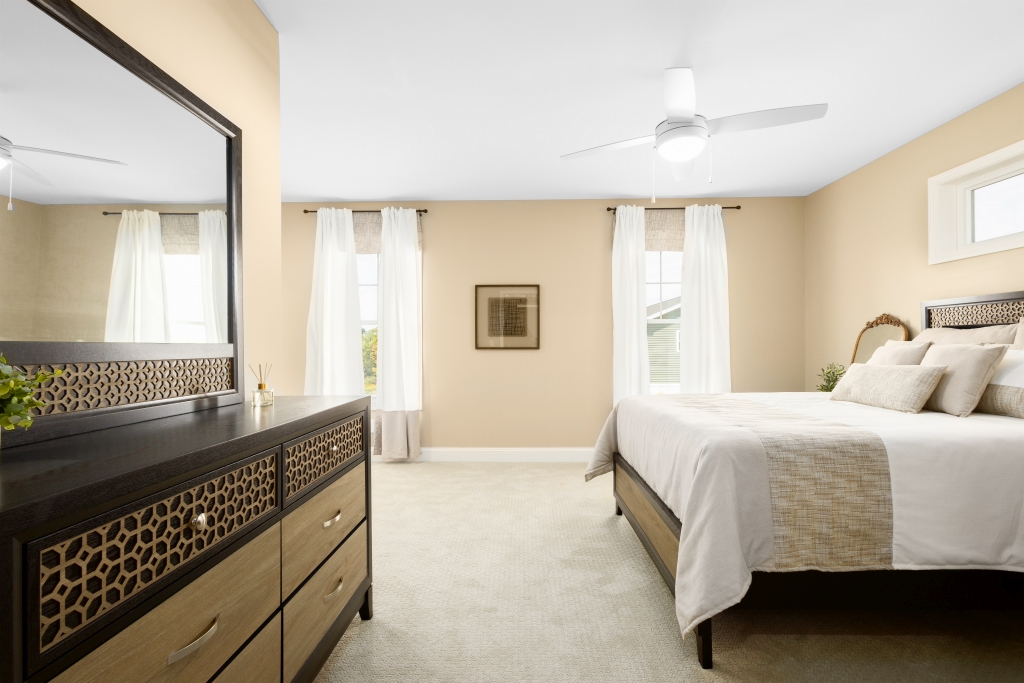 A serene bedroom with beige walls, a large bed with neutral linens, two tall windows with sheer curtains, a dresser with a mirror, and a ceiling fan.