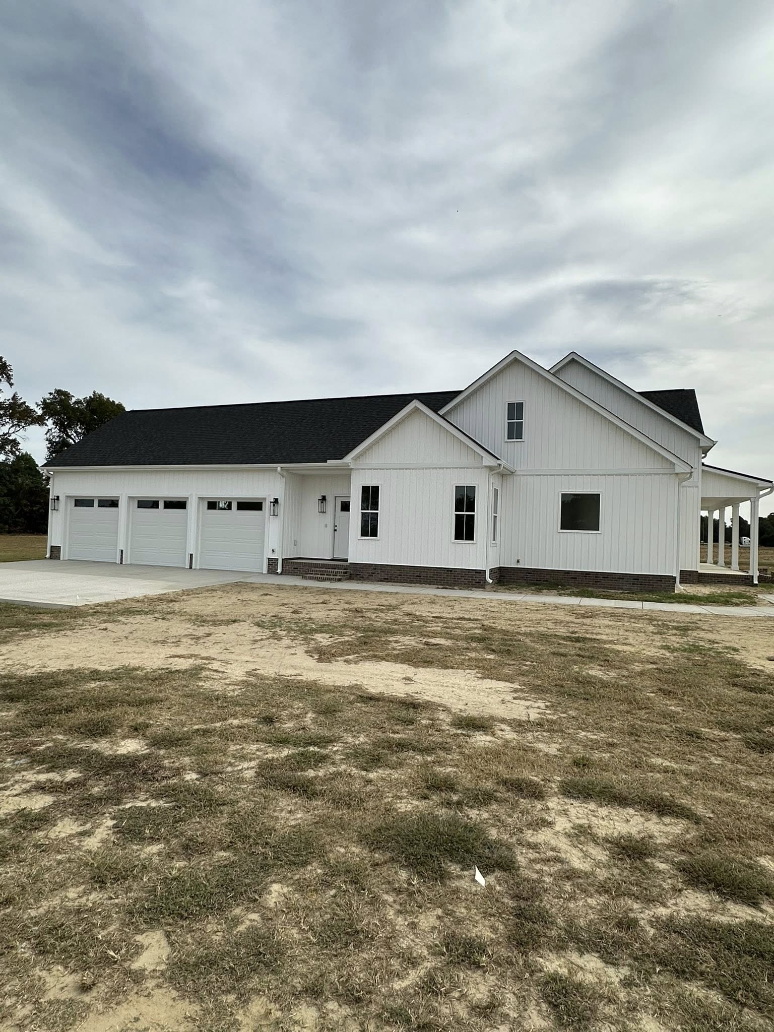 A modern white house with a black roof sits on a barren patch of land. It features multiple gables, large windows, and a three-car garage under a cloudy sky.