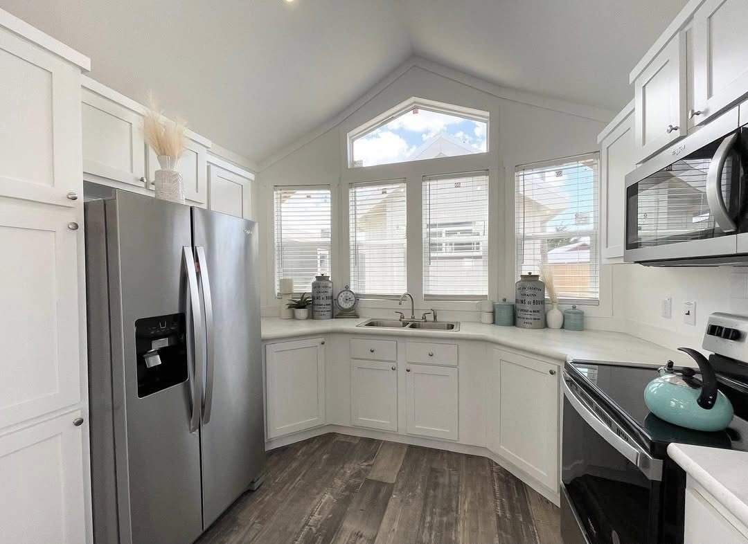 Bright kitchen with vaulted ceiling, large windows, and white cabinetry. Stainless steel appliances, including fridge and stove. Light wood floors add warmth.