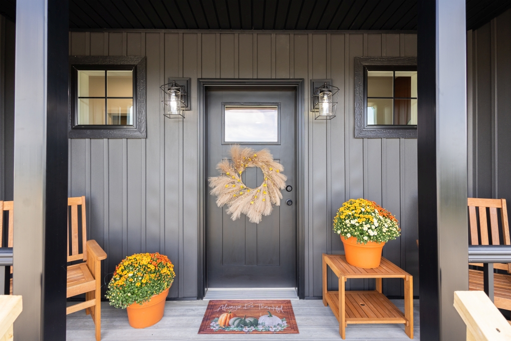 A cozy front porch with a black door adorned with a wheat wreath. Two potted flowers sit on wooden stands, framed by two wooden chairs, creating an inviting autumnal ambiance.