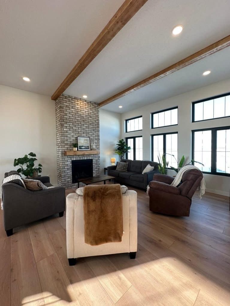 Chic living room with a brick fireplace, wooden beams, and large windows. Cozy sofas and armchair enhance the warm, inviting atmosphere.