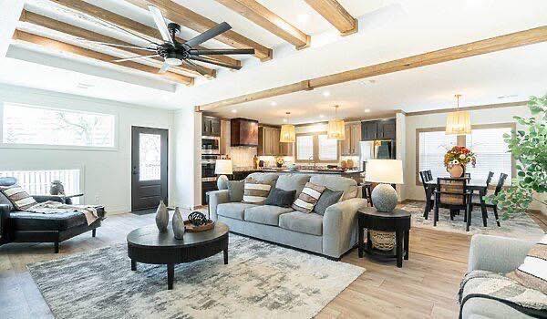 A spacious, modern living room with wooden beams, light gray sofa, and round coffee table. Open kitchen with dark cabinets and cozy dining area. Bright and inviting.