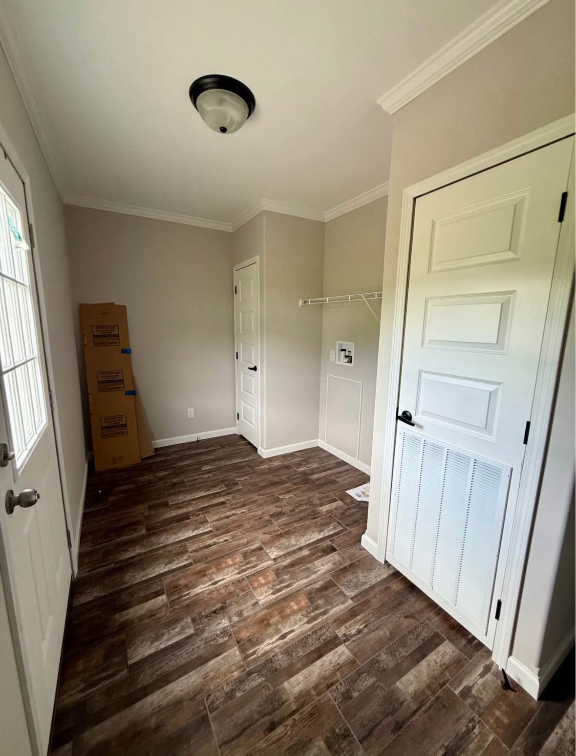A small laundry room with wood-textured tile flooring, light gray walls, and a ceiling light. Cardboard boxes sit in the corner, and a white door is ajar.