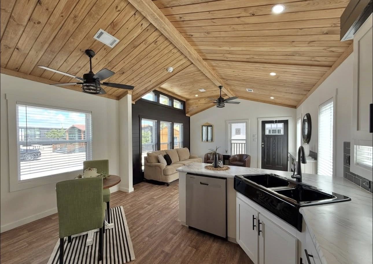 Spacious living room and kitchen with wooden ceiling, modern furniture, green chairs by a small round table, and large windows letting in natural light.