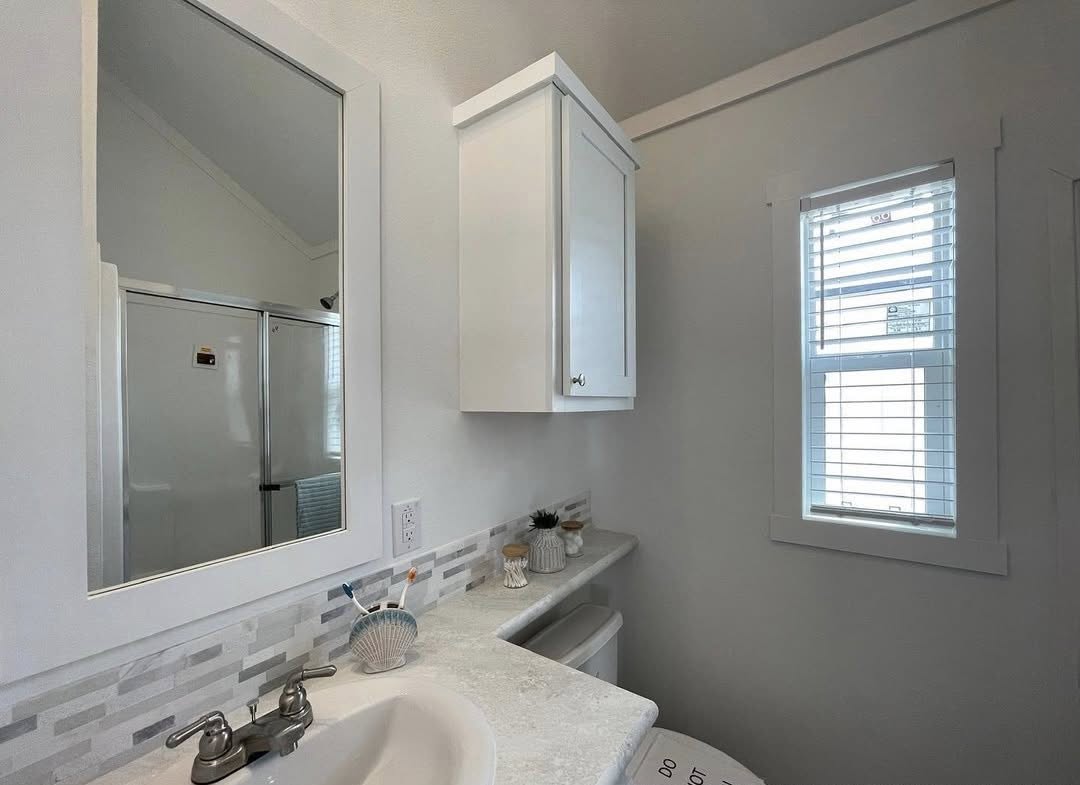 Bright bathroom with a white vanity, a mirror above, and a tiled backsplash. A shower with sliding doors is visible, and light streams through blinds.