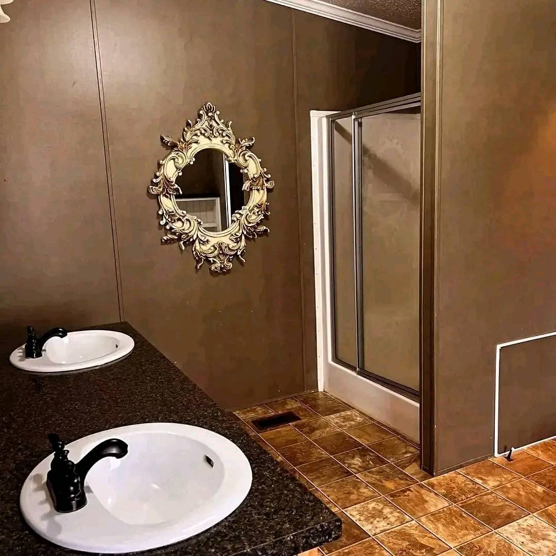 A bathroom with dark brown walls features an ornate oval mirror above dual sinks with black faucets on a granite countertop. A glass shower is in the corner.