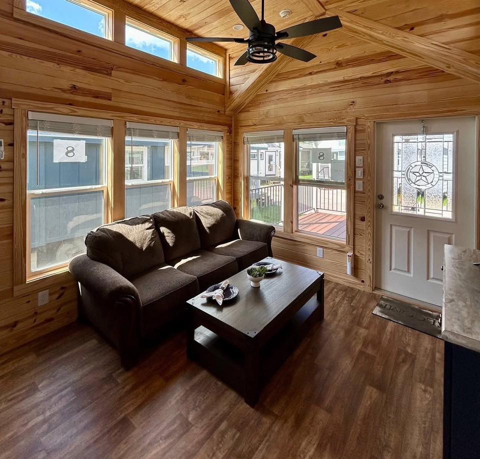 Cozy wood-paneled living room with large windows, brown sofa, wooden coffee table, and ceiling fan. Bright natural light creates a warm ambiance.