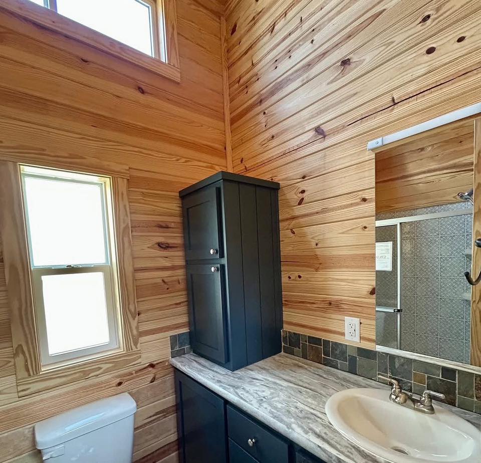 Cozy bathroom with wood-paneled walls, a small window, dark cabinet, marble countertop, and a white sink. Rustic and serene atmosphere.