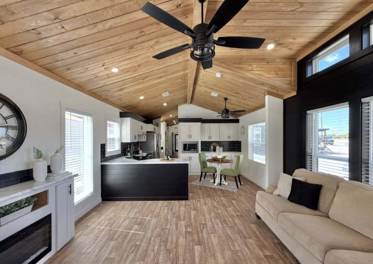 Spacious modern living area with wooden vaulted ceiling, open kitchen, dining nook, and beige sofa. Features ceiling fans, large windows, warm tones.